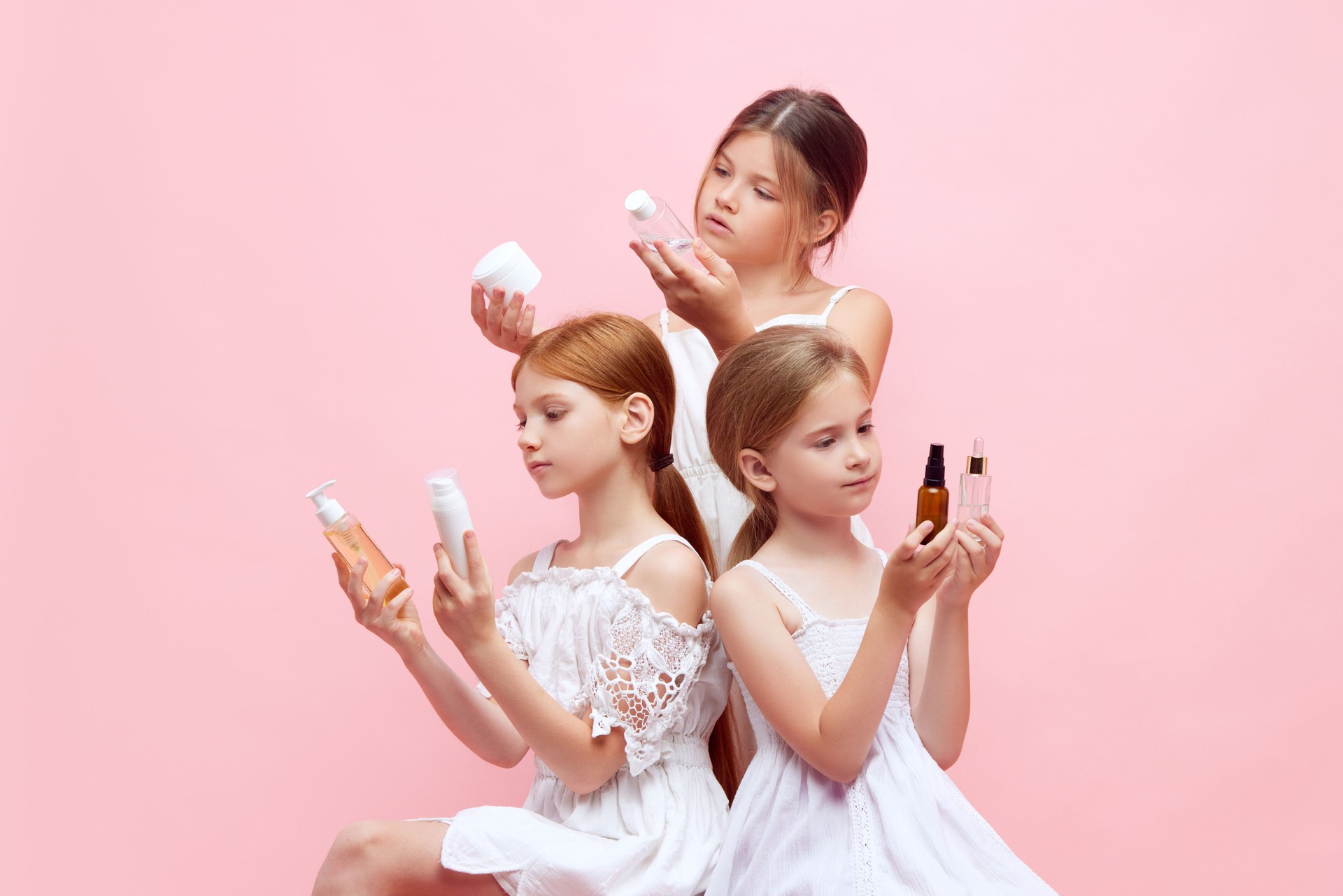 Beautiful, cute, little girls, children in white dresses design with organic cosmetological face products for kids skin care against pink studio background